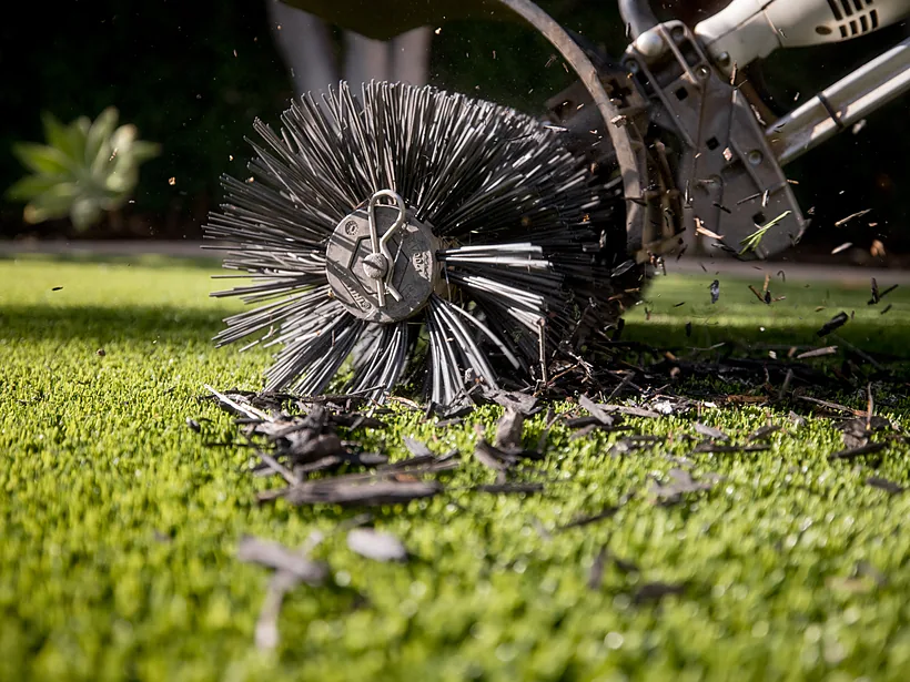 Power brush grooming embedded debris out of artificial turf fibers