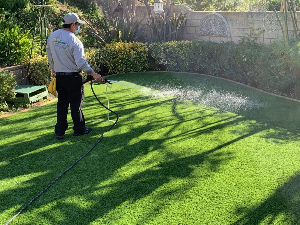 Technician applying TurFresh cleaner to artificial turf in a backyard, showcasing its eco-friendly, non-toxic formula that eliminates odors and is safe for kids and pets.