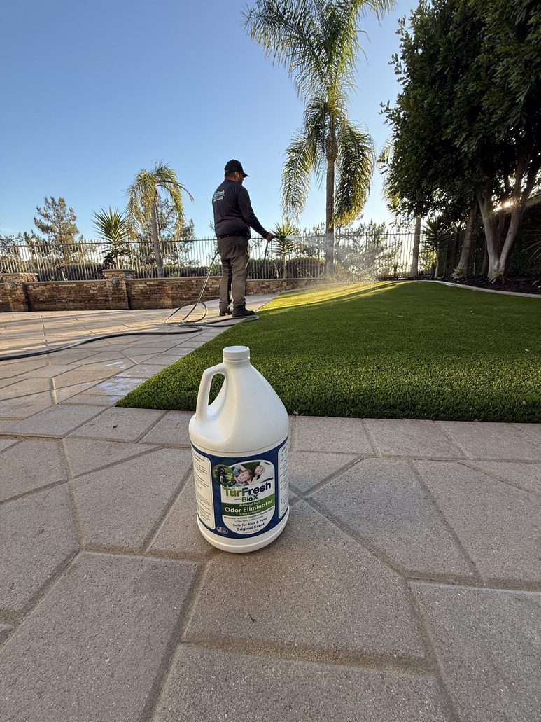 "Technician using TurFresh turf cleaner on artificial grass with a TurFresh BioTurf odor eliminator bottle in the foreground, showcasing its eco-friendly, pet-safe, and odor-removing benefits."